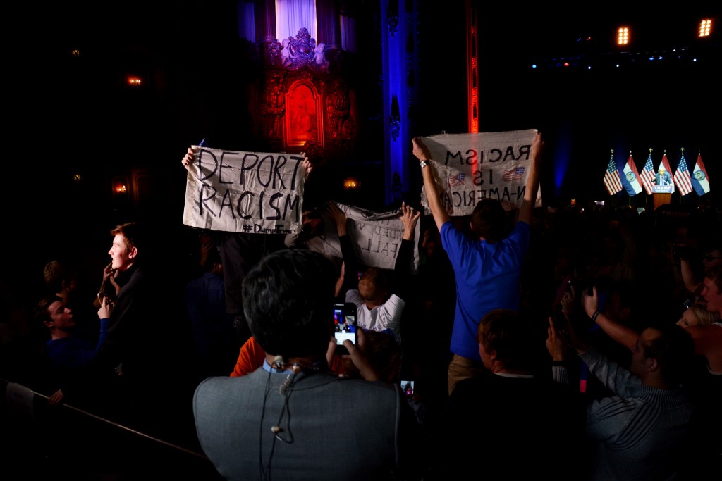 A group of protesters stand up and wave homemade flags shortly before being removed by police. Many protesters criticized Trump’s alleged racism and disdain of various groups. Photo by Andrew Hartnett