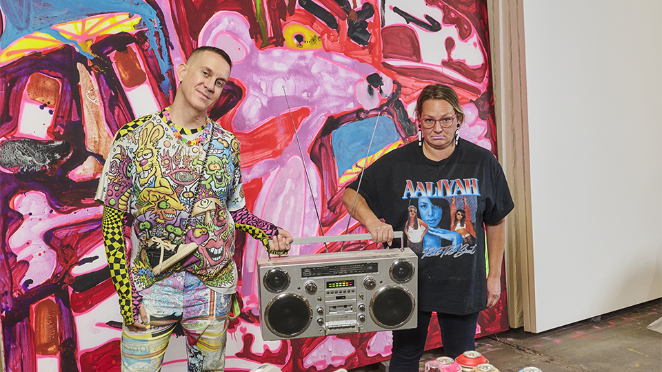 Man and woman holding a boom box between them, standing in front of a large pink panther painting.